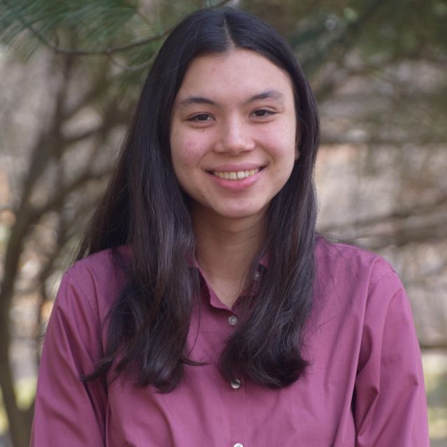 Headshot of Darcy standing in front of a pine tree. She is wearing a pink button-up shirt