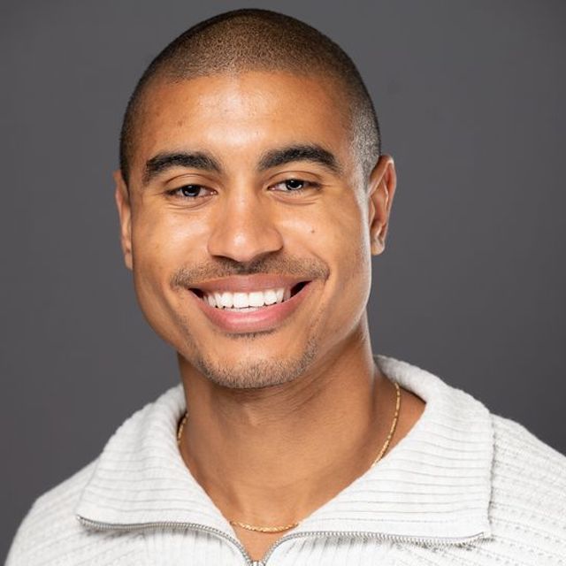 Headshot of Bryce Barbee, he is wearing a white sweater, a necklace and there is a grey background